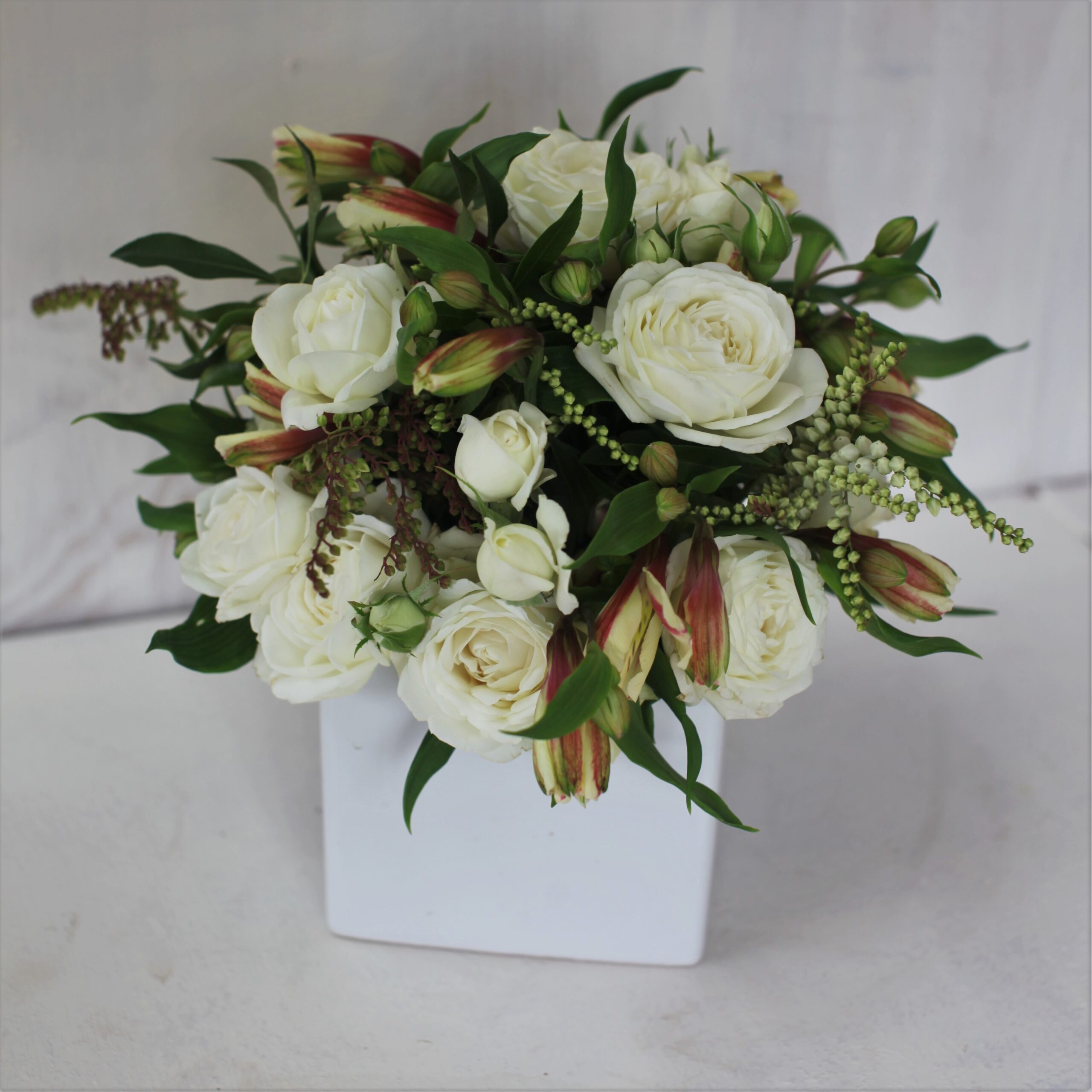 A square white ceramic container is filled with a posy of flowers including alstroemeria and roses.